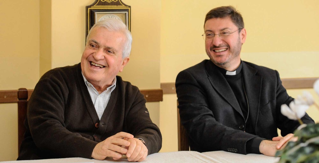 Deux prêtres en conversation, souriants, autour d’une table.